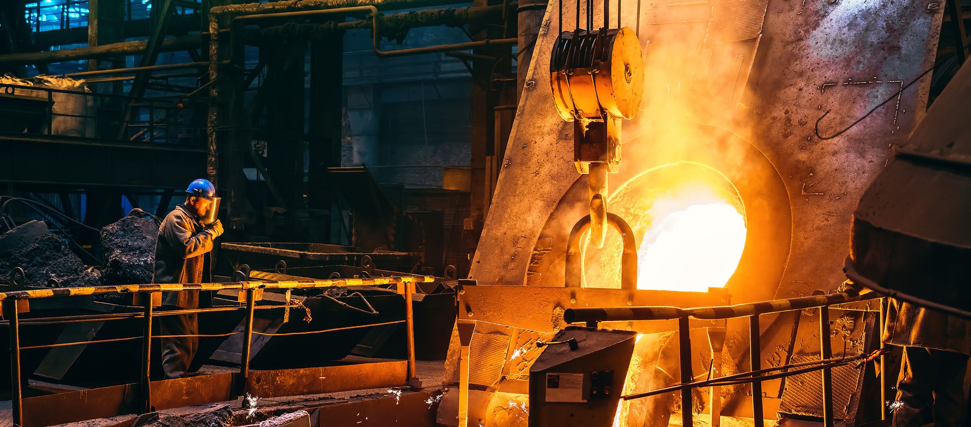 Molten steel pouring from a ladle inside a mill melt shop as a worker shields their face from the heat